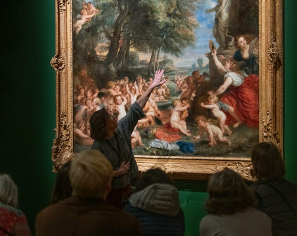 Vuxna personer sitter och tittar på en målning på en museivägg. En kvinna står upp oh pekar på målningen, hon ser ut att berätta om den.