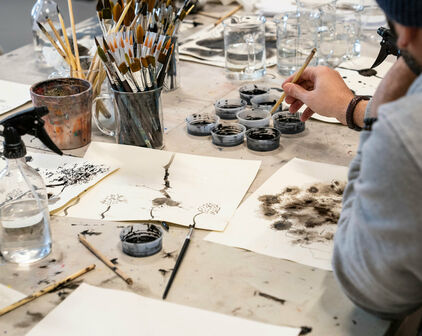 Papper, penslar, pennor och annat materiel för att teckna. En person sitter vid ett bord och tecknar med tusch.